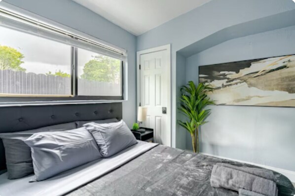 A bedroom with a bed, a window, a door, and a painting on the wall.