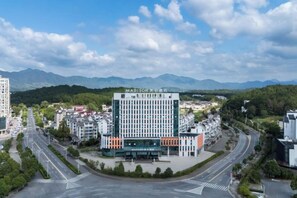 A multi-story hotel with a prominent sign, surrounded by a road and greenery, with mountains in the background. - Madison Hotel Huangshan Scenic Area North Gate (Huangshan City)