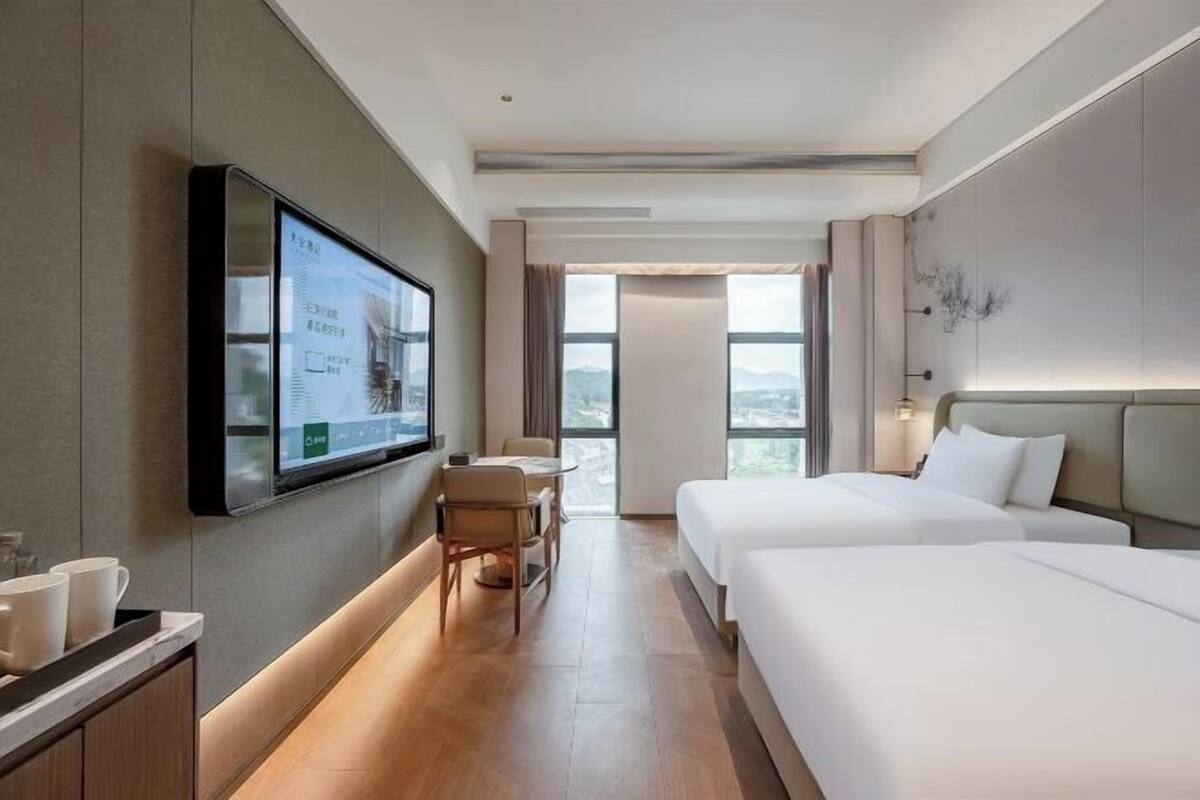 Ein Hotelzimmer mit Fernseher an der Wand, einem Schreibtisch mit Stuhl, zwei Betten und Blick aus dem Fenster.