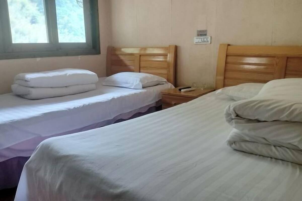 Ein Zimmer mit zwei Einzelbetten, beide mit Holz Kopfteilen und weißer Bettwäsche. Am linken Rand befindet sich ein Fenster und an der Wand eine Steckdose.