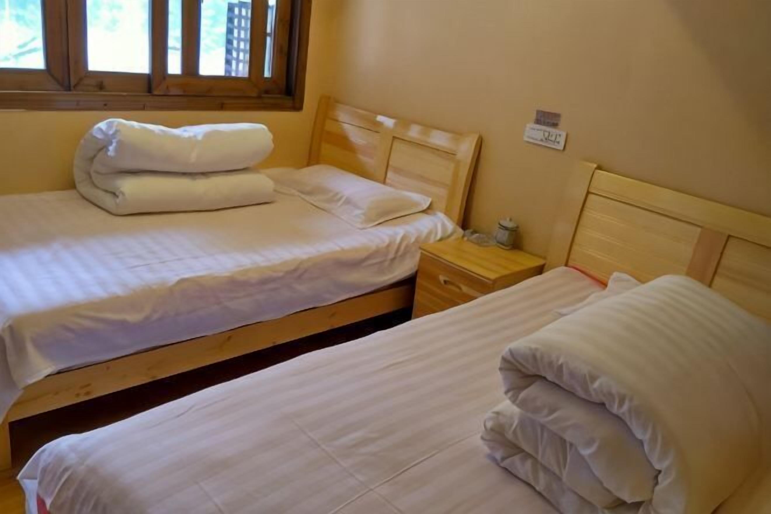 Una habitación con dos camas individuales, cabeceros de madera, una mesita de noche con lámpara y una ventana con persianas.