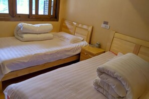 A room with two single beds, wooden headboards, a nightstand with a lamp, and a window with blinds.