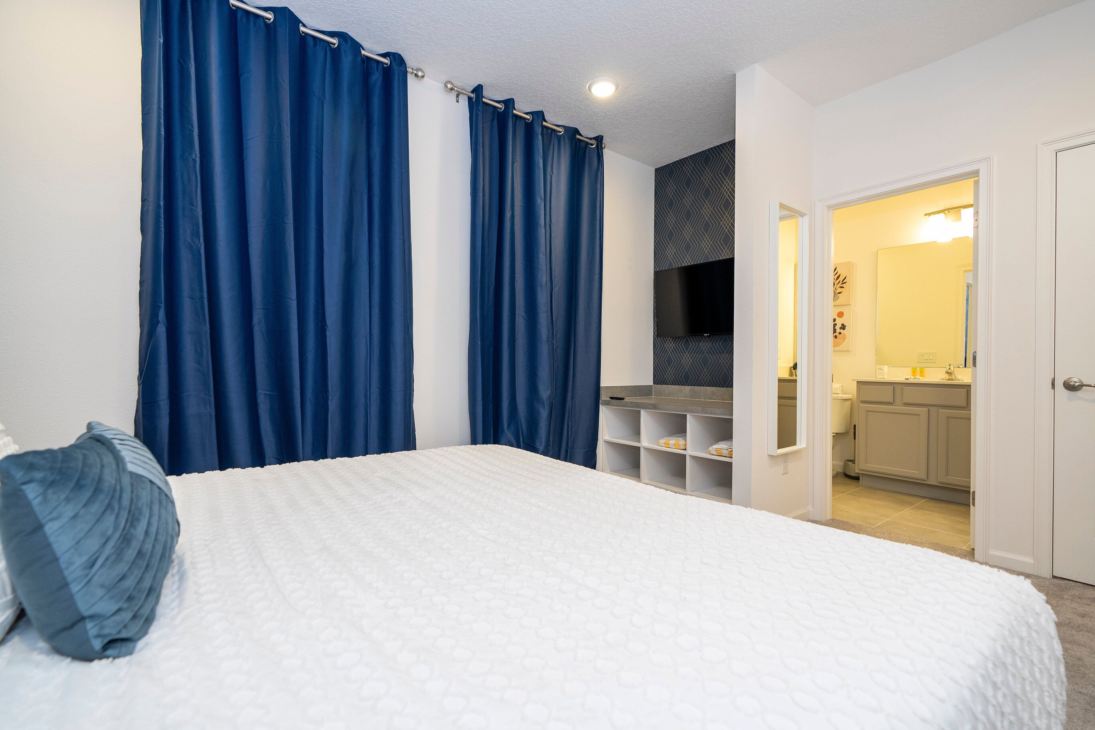 Ein Schlafzimmer mit Bett, blauen Vorhängen, einem weißen Kopfteil und einem fest installierten Fernseher.