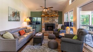 A cozy living room with a stone fireplace, comfortable seating, and a large window offering a view of the outdoors. - Fairway Gem at Spruce Pine - EV charge port, hot tub, fishing access (Spruce Pine)