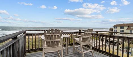 Een houten terras met twee Adirondack-stoelen met uitzicht op een strand en een gebouw.