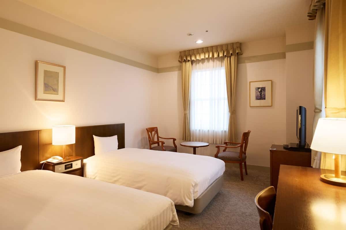 Ein Hotelzimmer mit zwei Betten, einem Schreibtisch, einem Stuhl, einem kleinen Tisch und einem Fernseher.