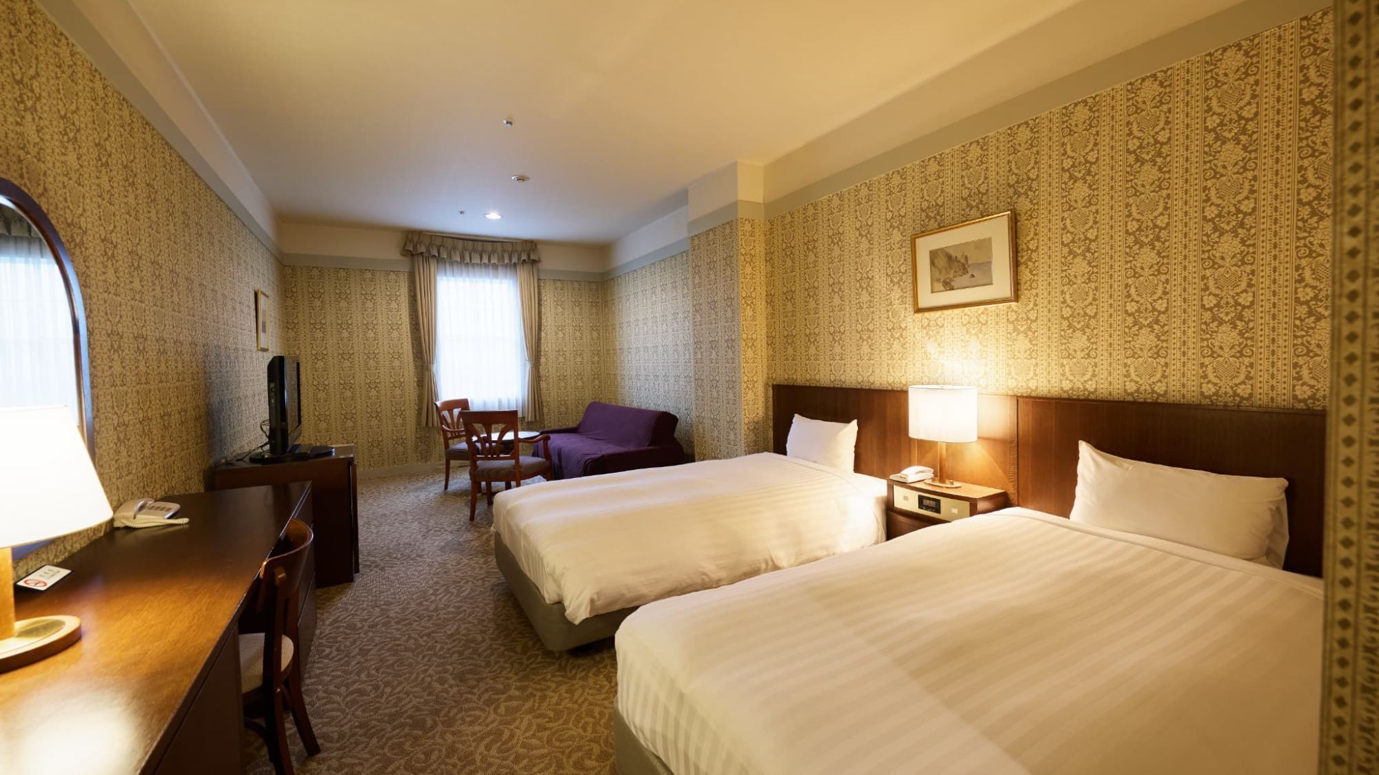 Ein Hotelzimmer mit zwei Betten, einem Schreibtisch, einem Stuhl, einem Fernseher und einem gerahmten Bild an der Wand.