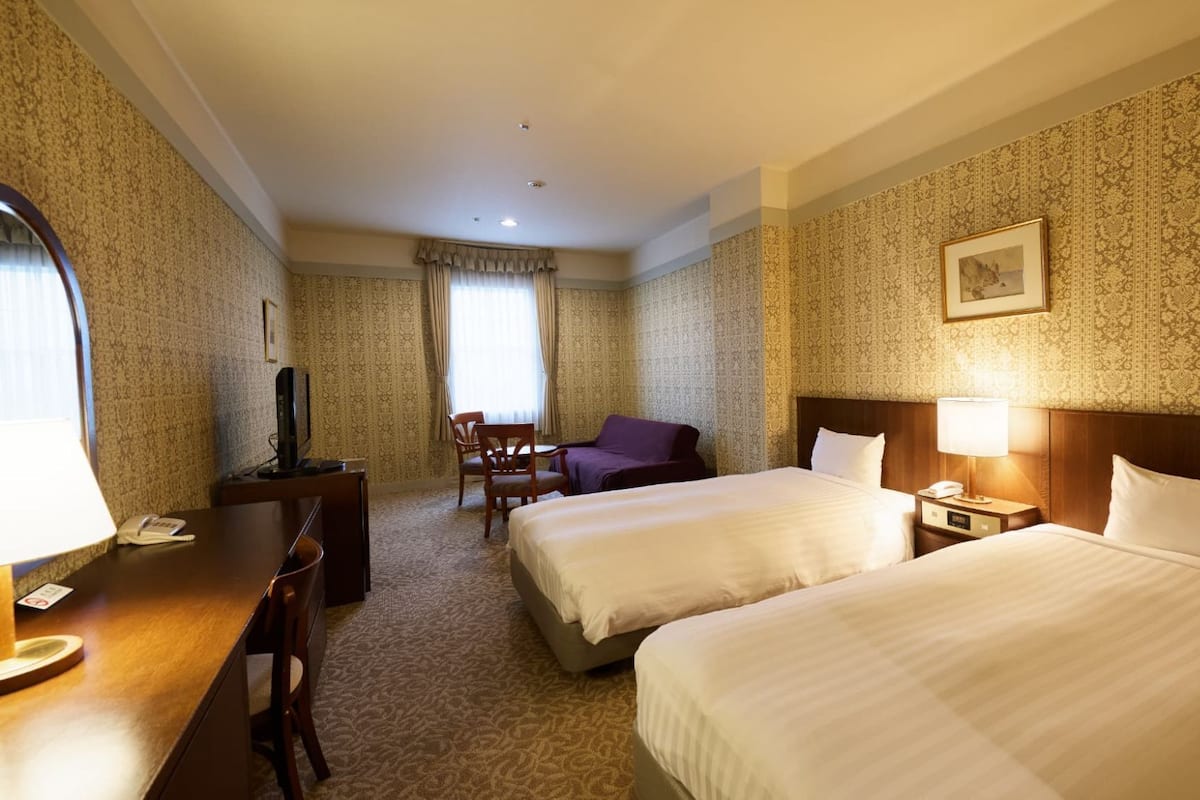 Ein Hotelzimmer mit zwei Betten, einem Schreibtisch, einem Stuhl, einem Fernseher und einem gerahmten Bild an der Wand.