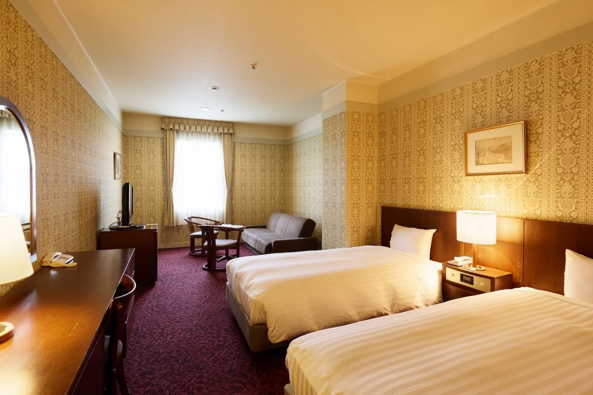 Ein Hotelzimmer mit zwei Betten, einem Schreibtisch mit Lampe, einem Sessel, einem Fernseher und einem Wandteppich mit Muster.