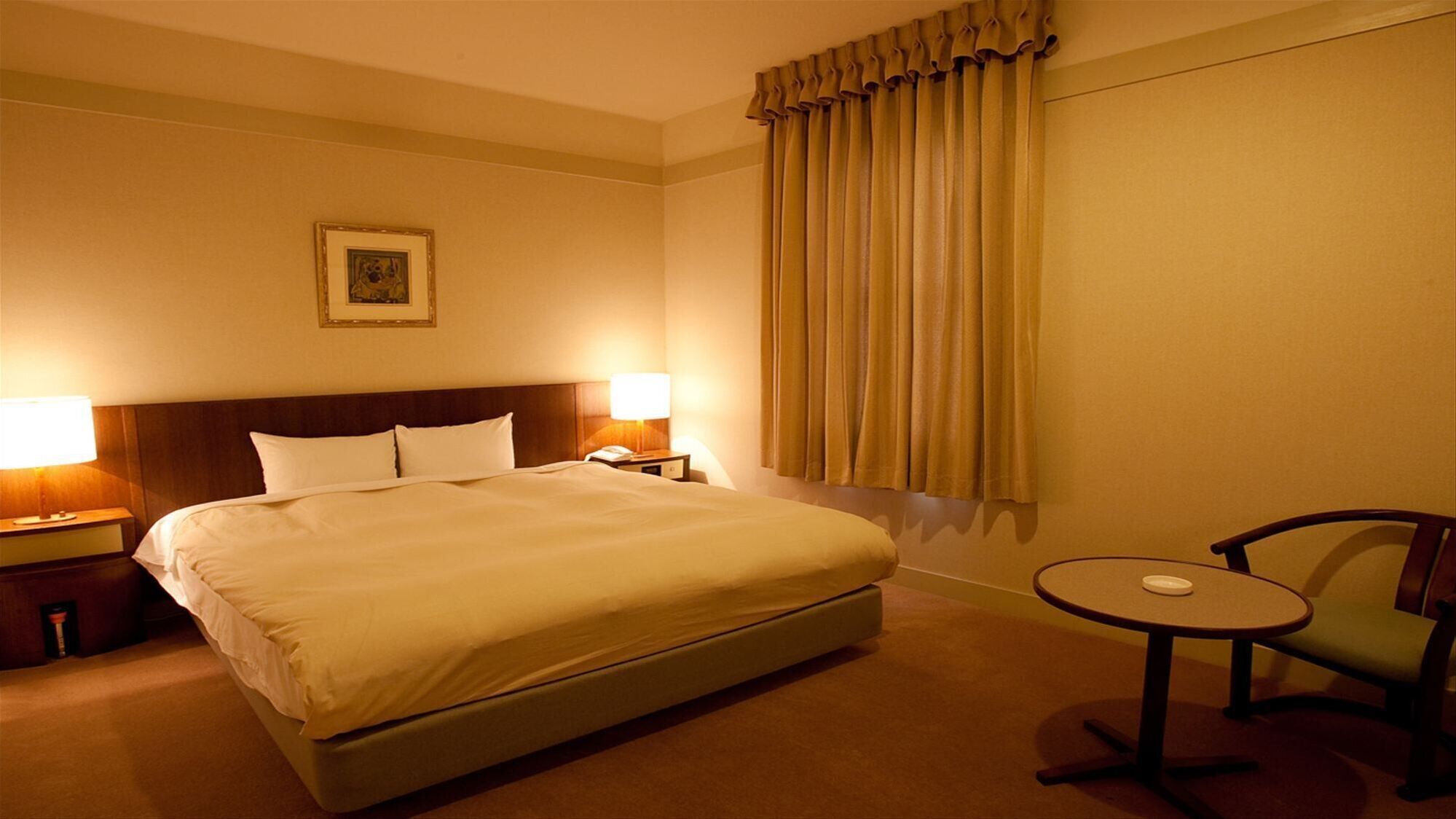 Ein Hotelzimmer mit einem großen Bett, zwei Nachttischlampen, einem kleinen Tisch, einem Sessel und einem Fenster mit Vorhängen.