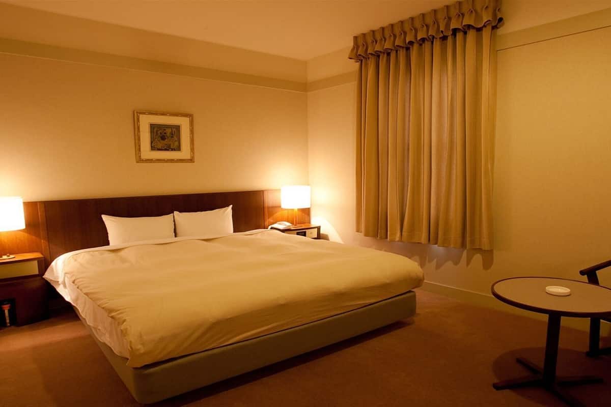 Ein Hotelzimmer mit einem großen Bett, zwei Nachttischlampen, einem kleinen Tisch, einem Sessel und einem Fenster mit Vorhängen.