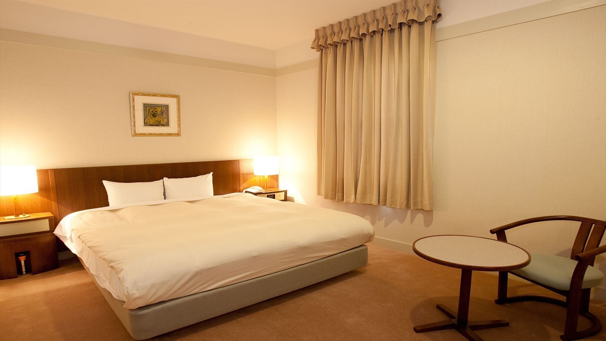 Ein Hotelzimmer mit einem großen Bett, zwei Nachttischlampen, einem kleinen Tisch, einem Stuhl und einem Fenster mit Vorhängen.