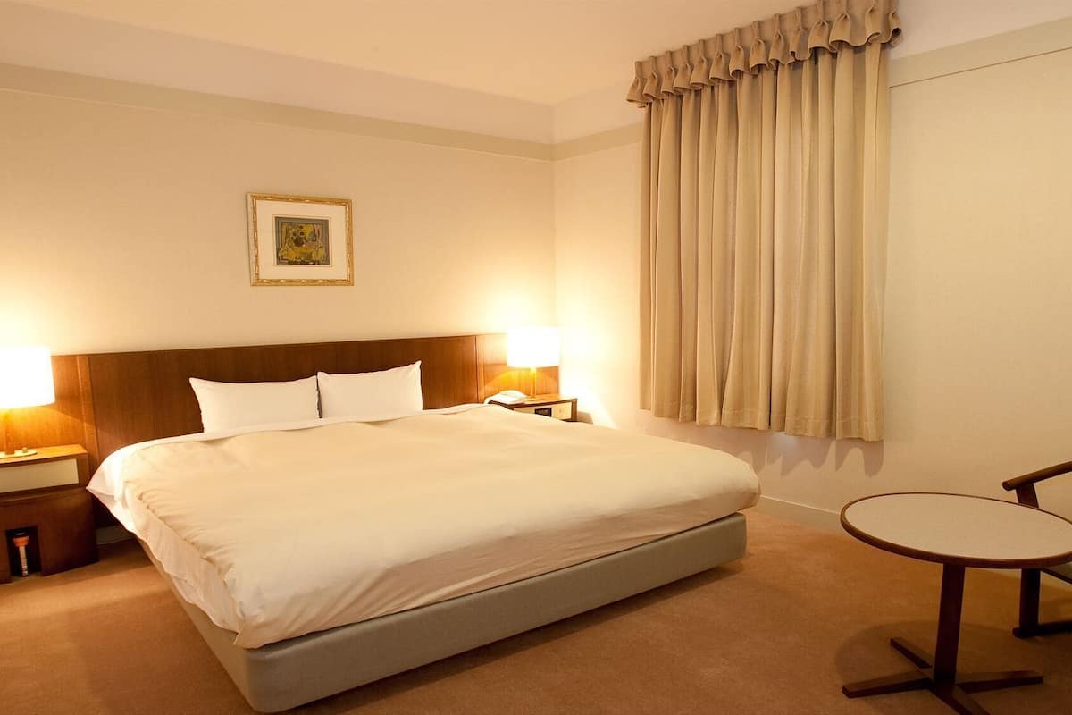 Ein Hotelzimmer mit einem großen Bett, zwei Nachttischlampen, einem kleinen Tisch, einem Stuhl und einem Fenster mit Vorhängen.