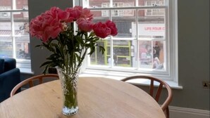 Une table ronde en bois, ornée d’un vase de fleurs roses, deux lampes suspendues et une vue sur un bâtiment en briques, visible à travers de grandes fenêtres.