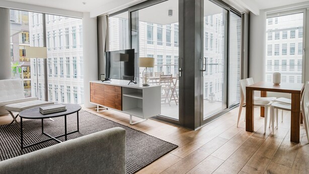 A modern living room with a large sliding glass door leading to a balcony, a flat-screen TV, a wooden cabinet, a dining table with chairs, and a sofa.