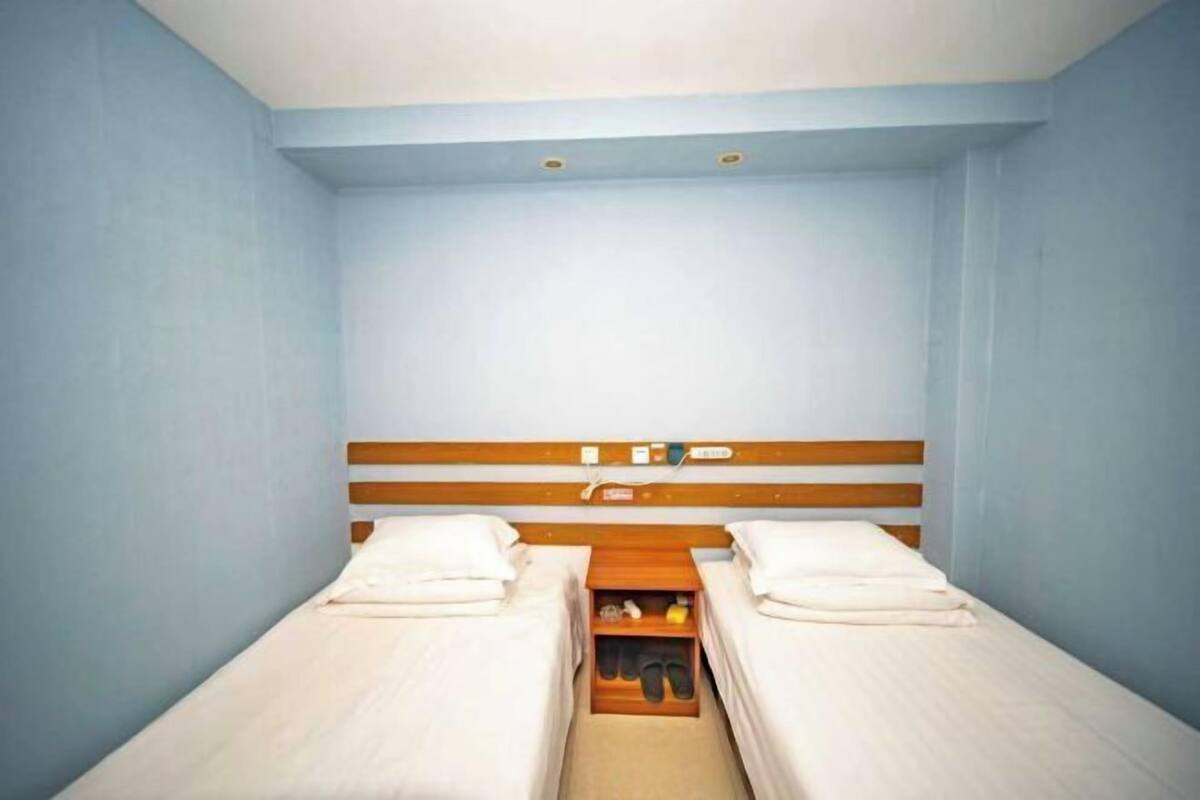 A small, single room with two beds, a wooden headboard, and a shelf with personal items.