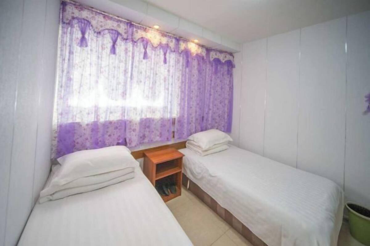 A small, clean room with two single beds, a wooden nightstand, and a window with purple curtains.