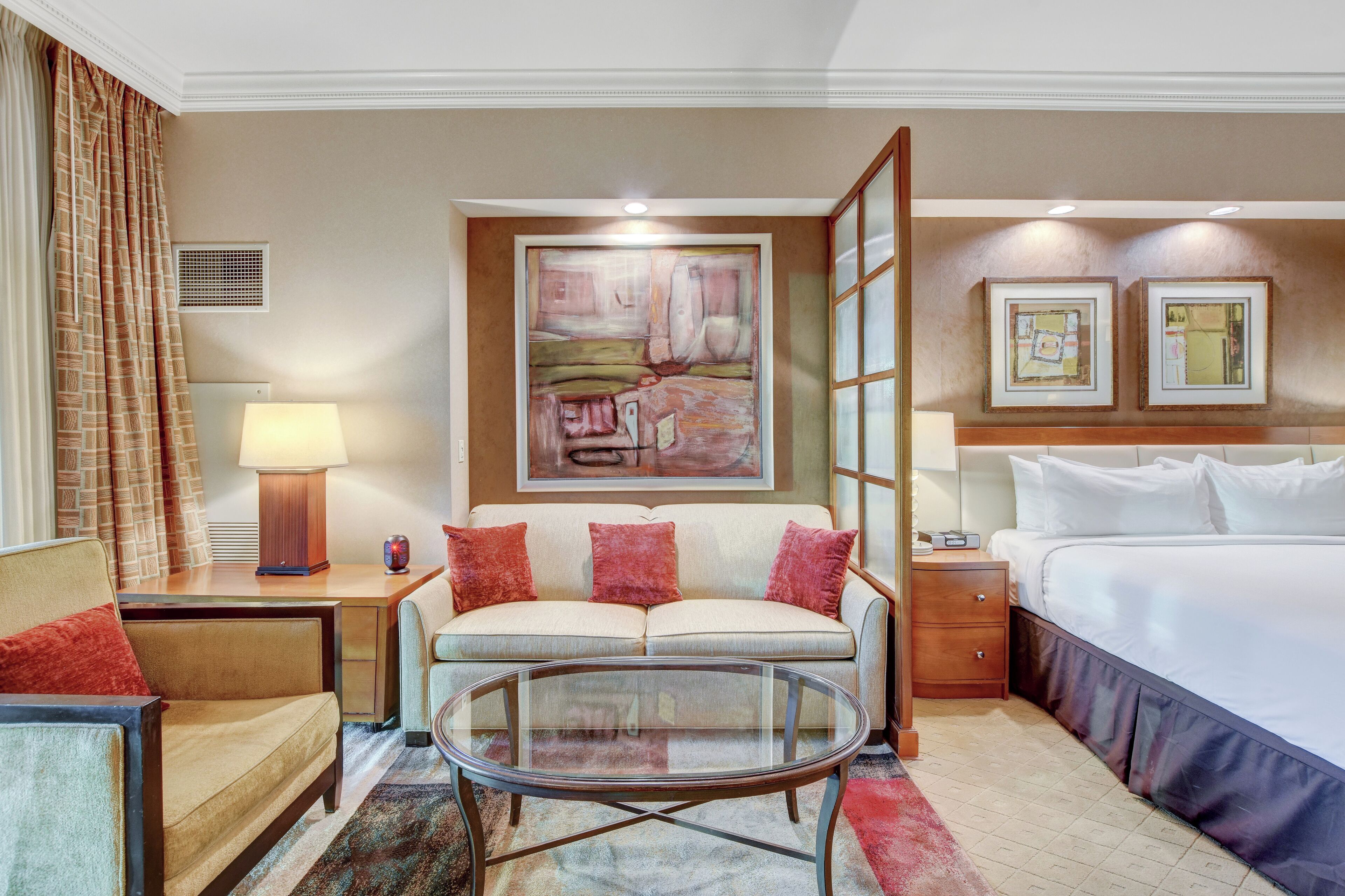 A hotel room with a sofa, a glass coffee table, a bed, a nightstand, a lamp, and a painting on the wall.
