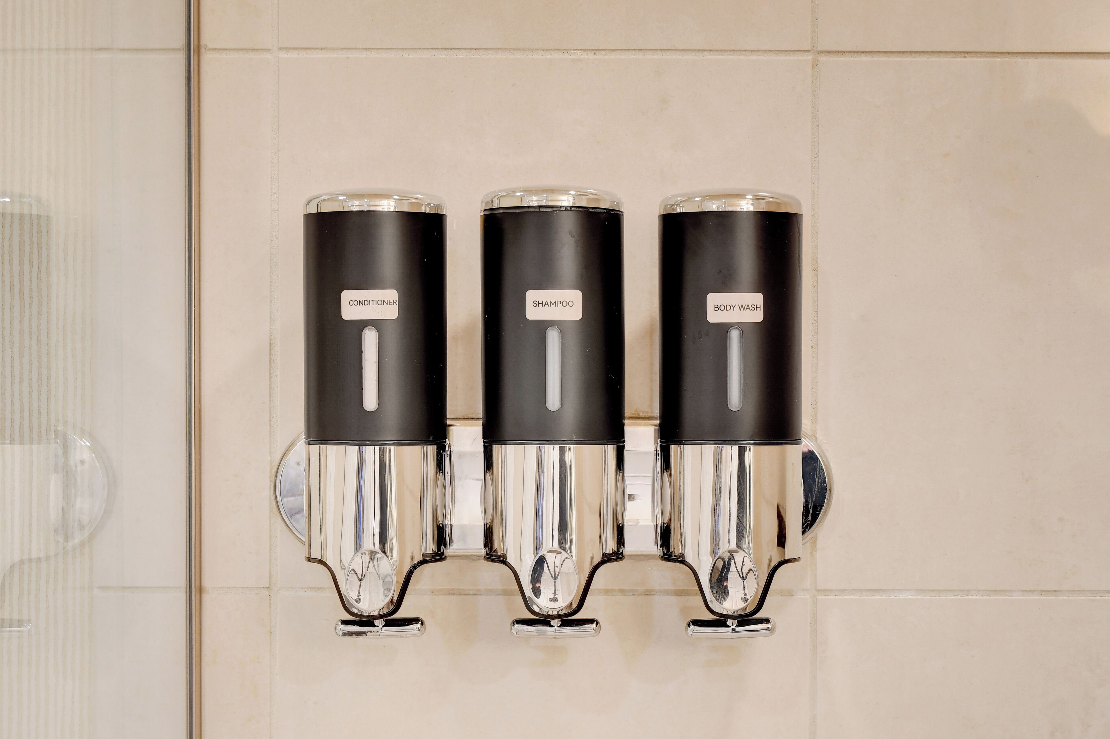 Dispensador fixado na parede com três compartimentos rotulados condicionador, champô e gel de banho.
