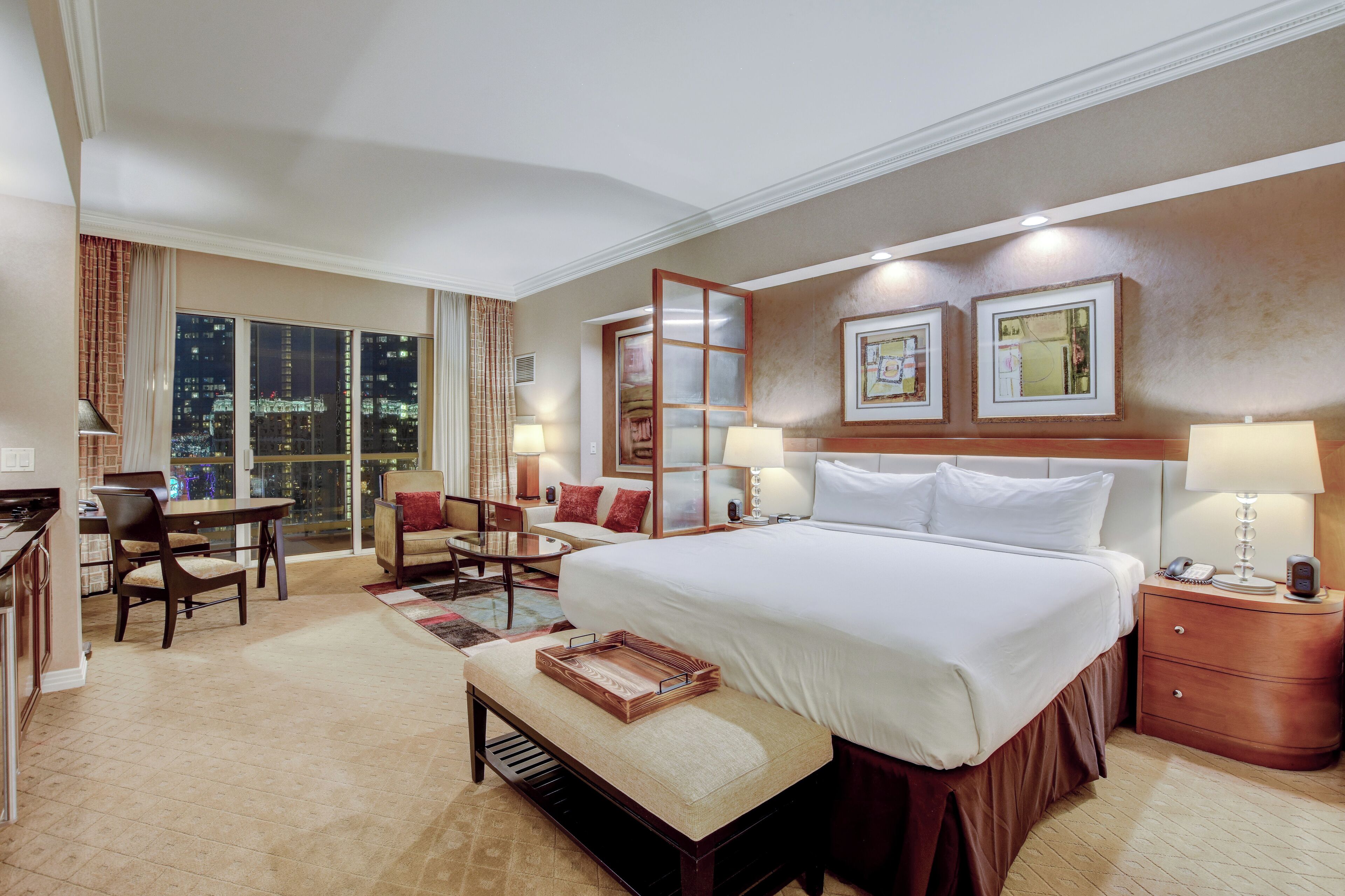 Ein Hotelzimmer mit einem großen Bett, einem Schreibtisch, einem Sessel, einem kleinen Tisch und Blick auf die beleuchtete Stadt.