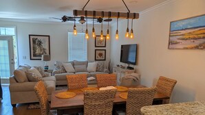 A living room with a sofa, a dining table with wicker chairs, a TV, and a ceiling light fixture. - Angler Villas 3 (Murrells Inlet)