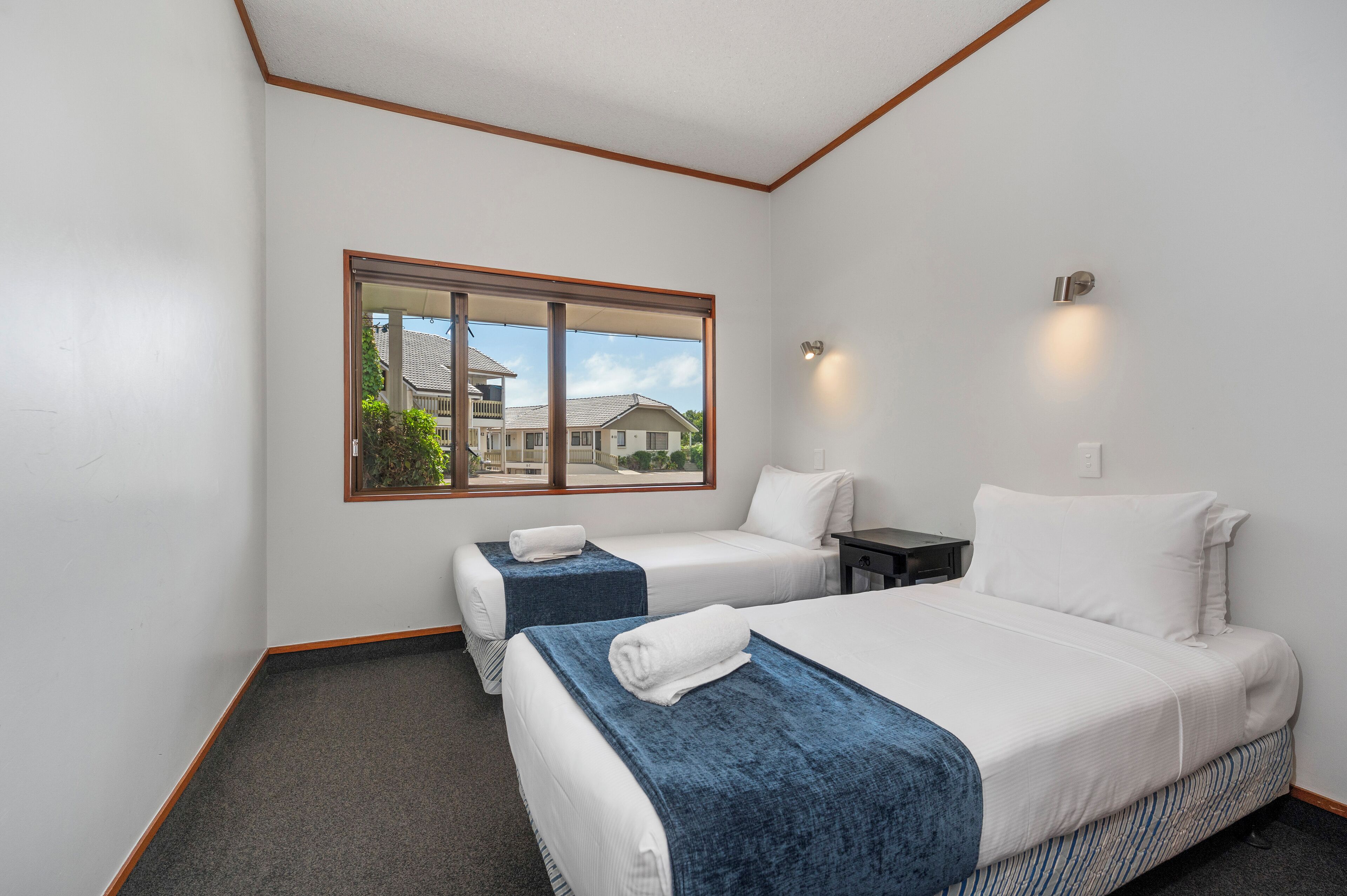 Habitación de hotel con dos camas, una ventana que da a vista de casas y una mesita con lámpara.