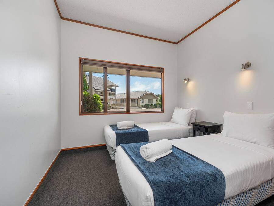 Habitación de hotel con dos camas, una ventana que da a vista de casas y una mesita con lámpara.
