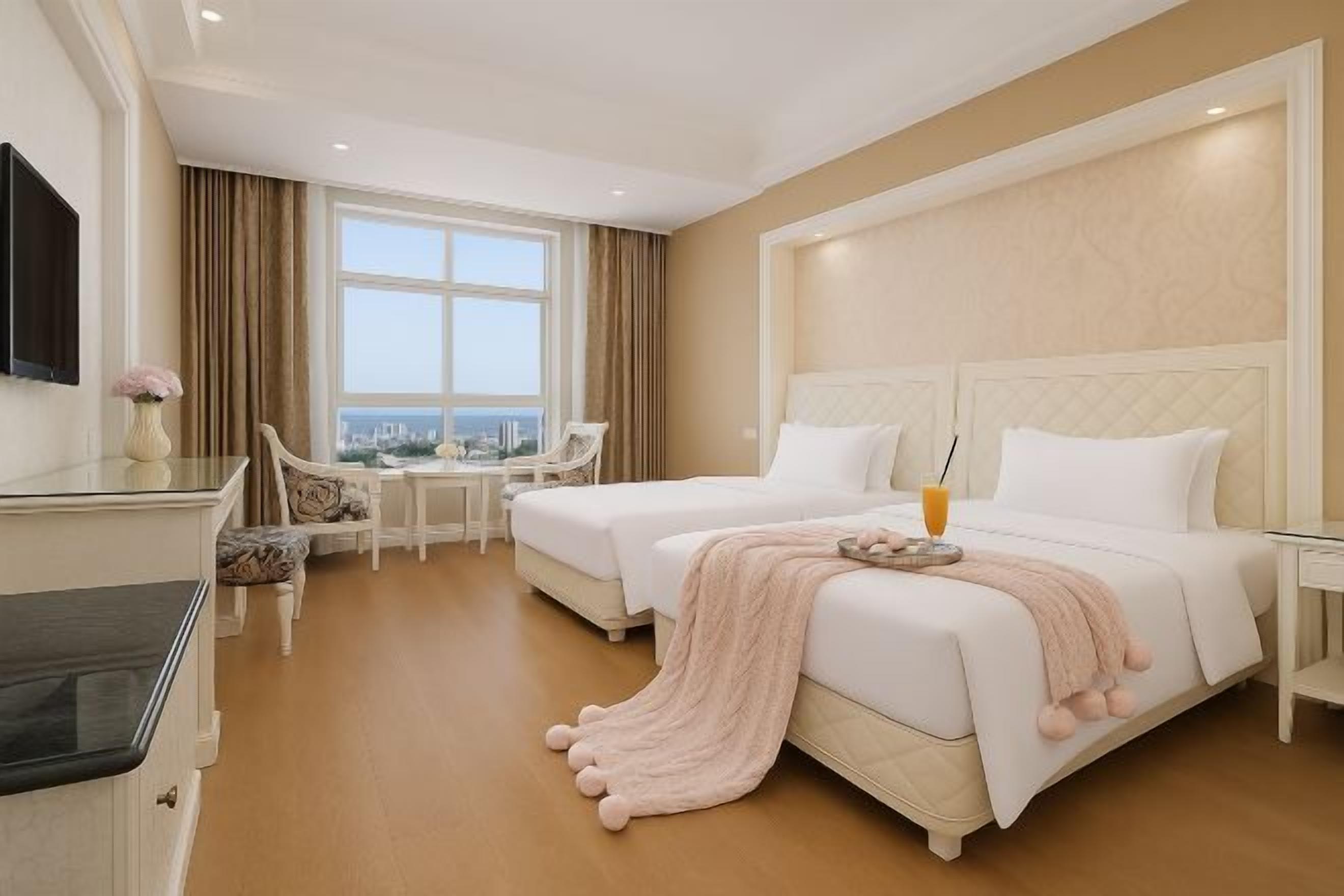 A hotel room with two beds, a desk, a chair, a TV, and a view of the city.