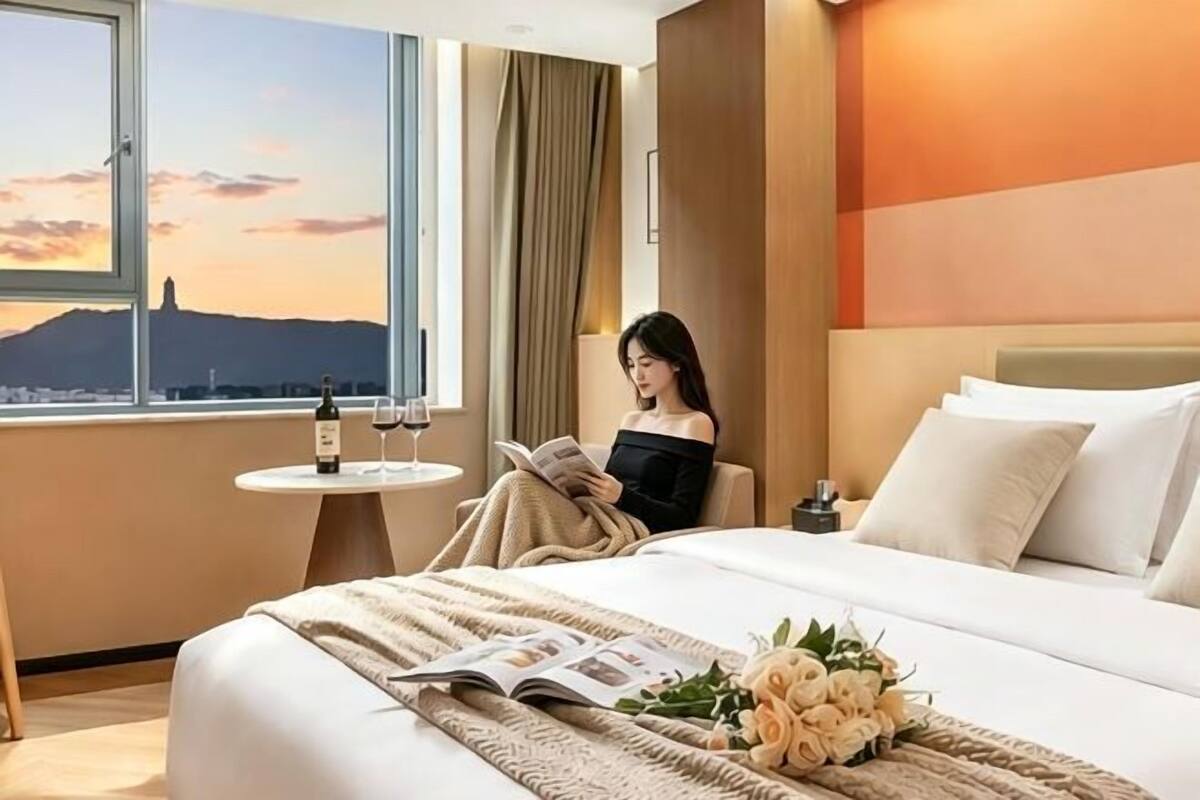 A hotel room with a bed, a window with a city view, a small table with a wine bottle and glass, and a bouquet of flowers.