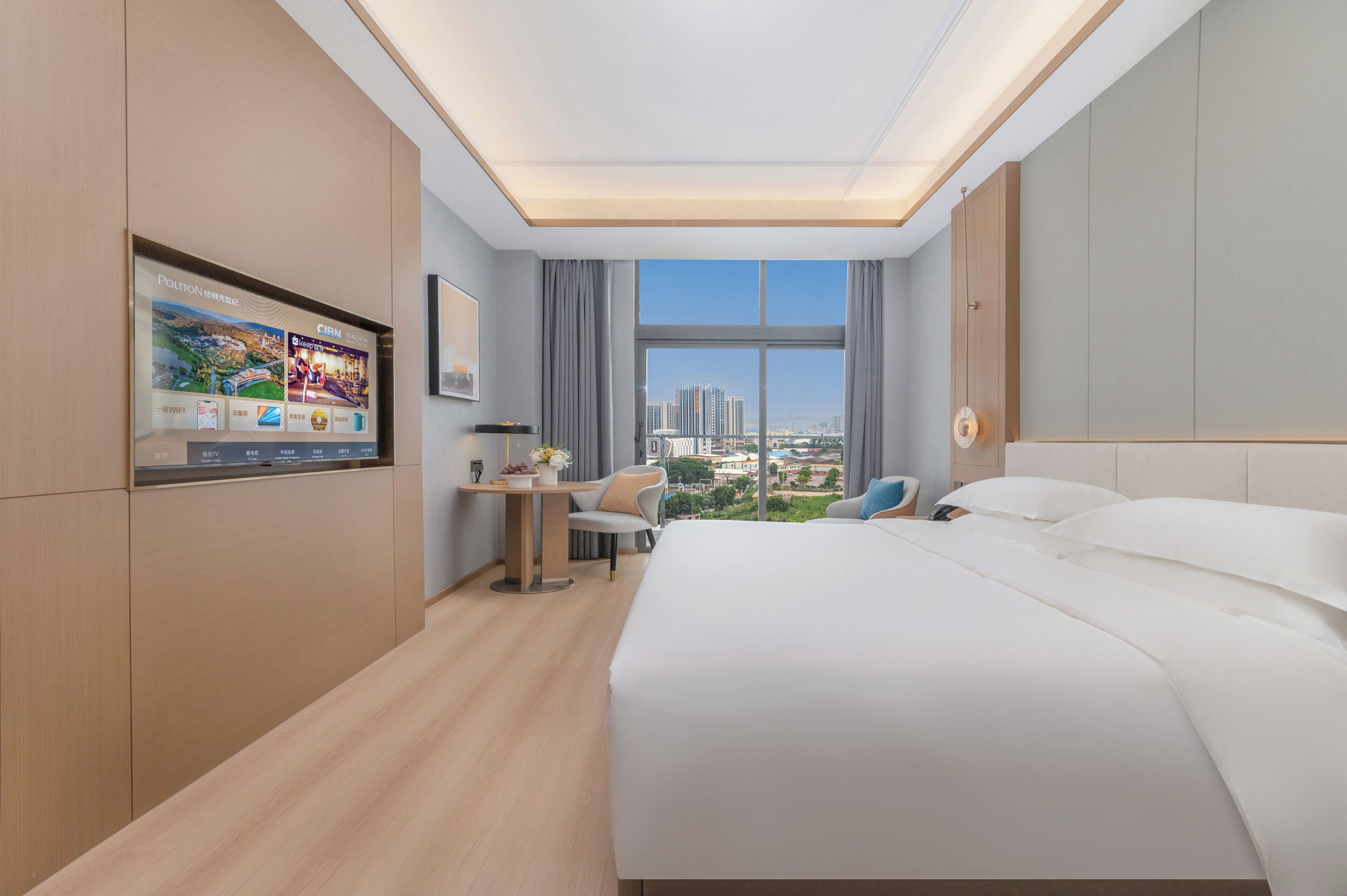 A hotel room with a large bed, a desk, a chair, a TV mounted on the wall, and a view of the city through the window.