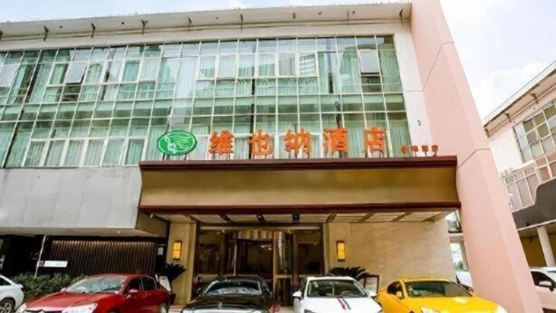 A hotel entrance with a glass facade, a green logo, and multiple cars parked in front. - Vienna Hotel (Suzhou)