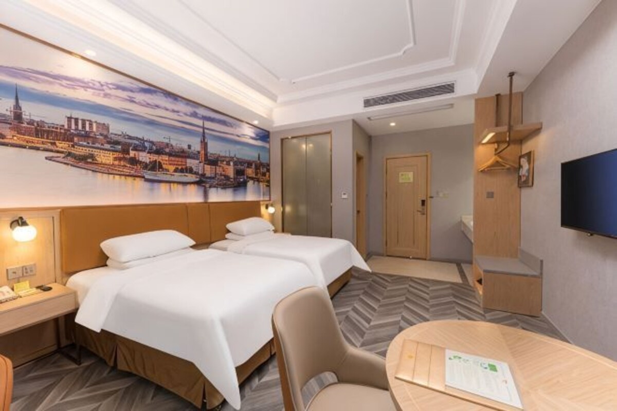 A hotel room with two beds, a desk, a chair, a TV, and a painting of a cityscape.