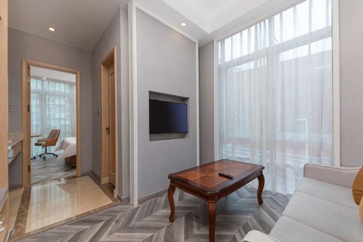 A modern living room with a sofa, a wooden coffee table, a mounted TV, and large windows.