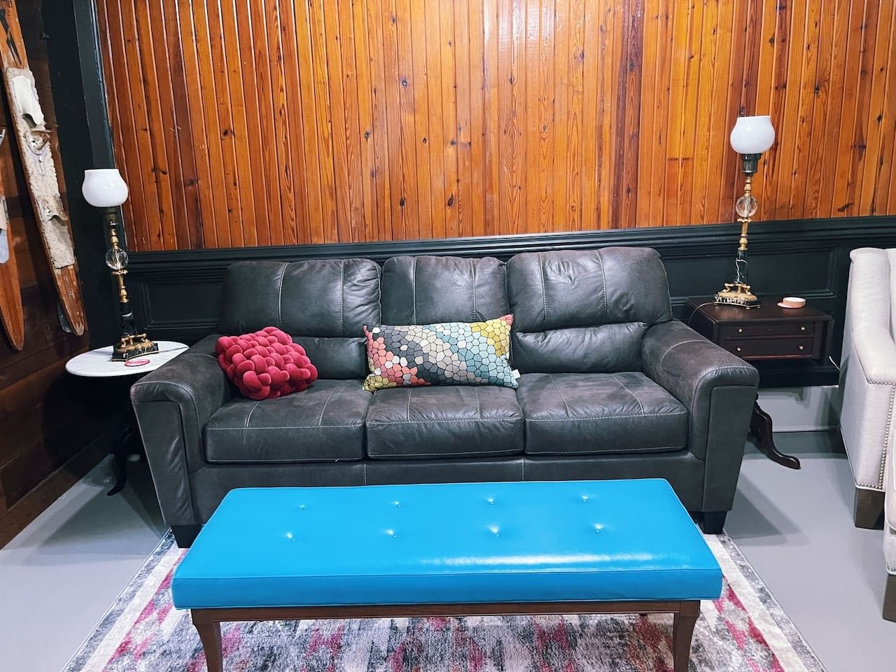 A room with a black leather sofa, a blue ottoman, and a wooden paneled wall.
