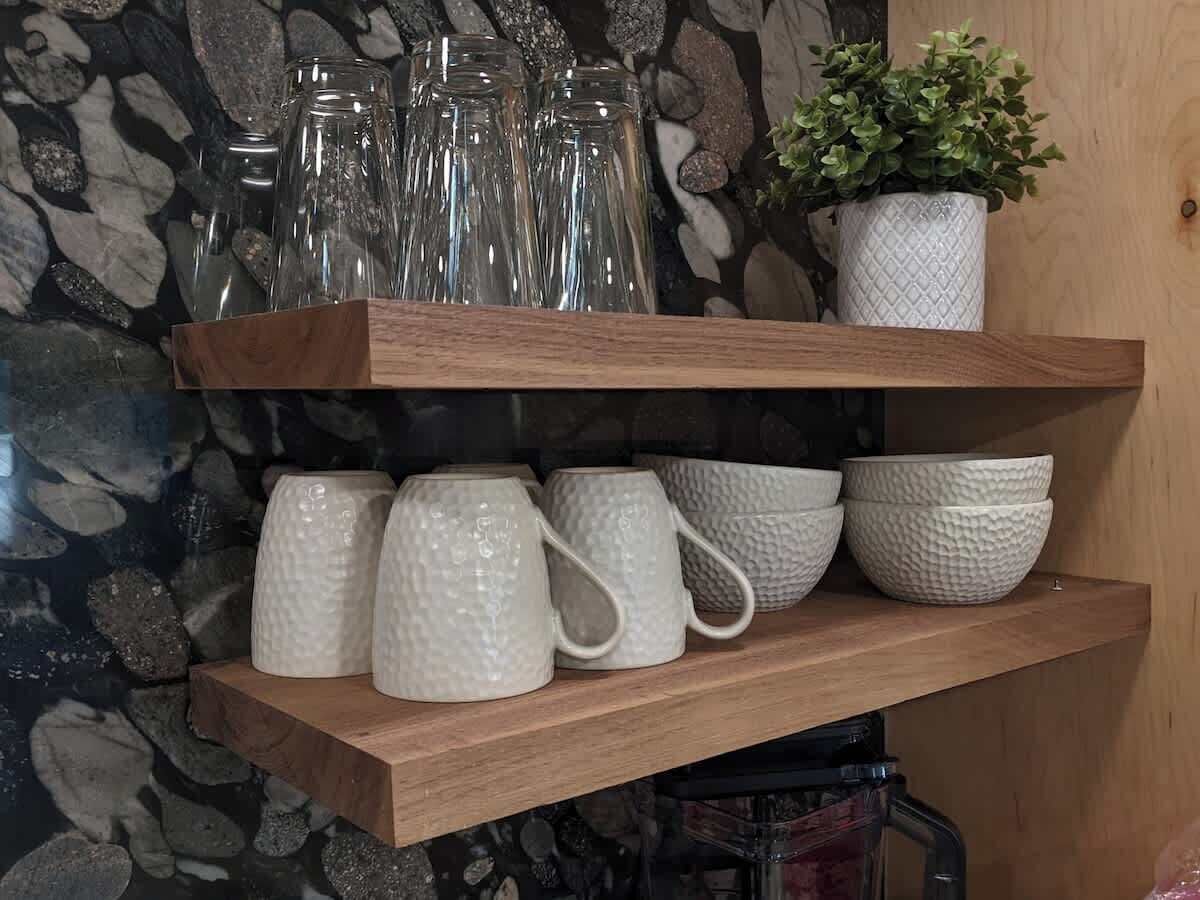 Ein Holzregal mit weißen, strukturierten Tassen und Schüsseln, einer Topfpflanze und klaren Glasflaschen.