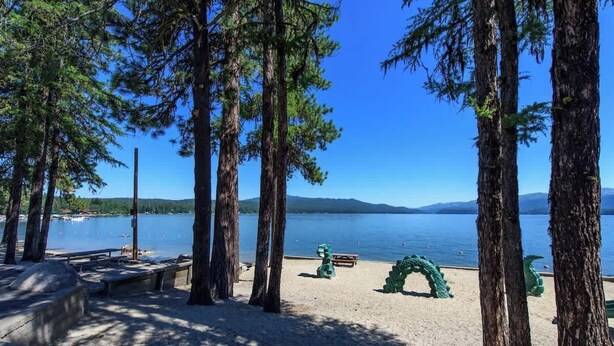 A lakeside park with tall trees, a sandy beach, and a clear view of the lake and mountains in the distance. - Updated Cabin HotTub Firepit Decks nr Shore Lodge (McCall)