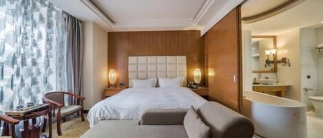 Habitación de hotel con cama, sofá, escritorio, silla, baño con bañera y un ventanal grande con cortinas.