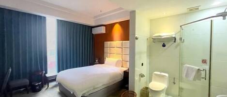 A hotel room with a bed, a desk, a chair, a bathroom with a toilet, and a shower.