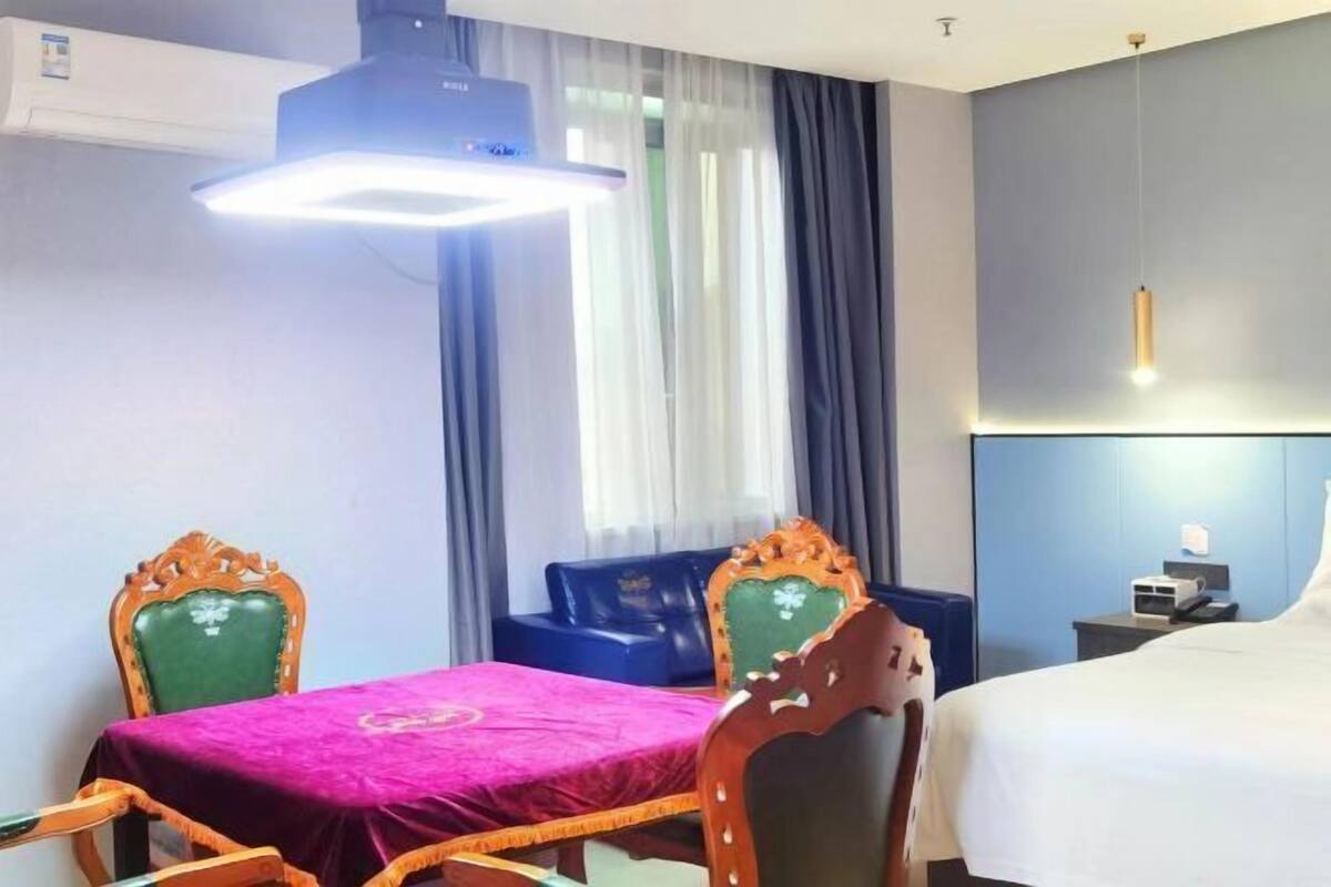 A hotel room with a bed, a desk, a chair, a table with a pink tablecloth, and a window with curtains.
