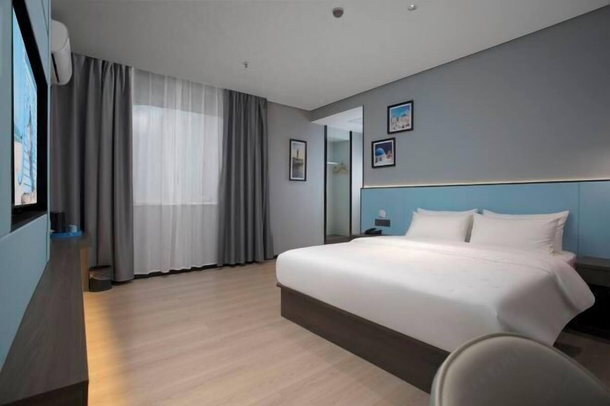 A modern hotel room with a large bed, a desk, a chair, and a window with curtains.