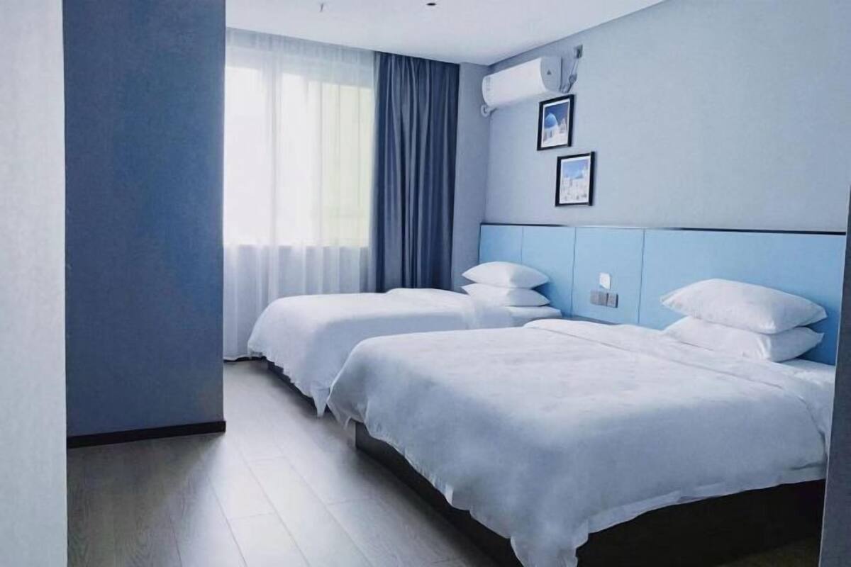 A hotel room with two beds, a large window with curtains, and a wall-mounted air conditioner.