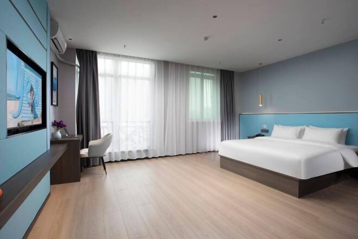 A modern hotel room with a large bed, a desk with a chair, a TV, and a large window with curtains.