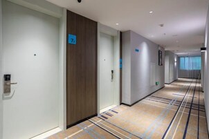 Pasillo de hotel con puertas numeradas, piso alfombrado y vista a una ventana con cortinas.