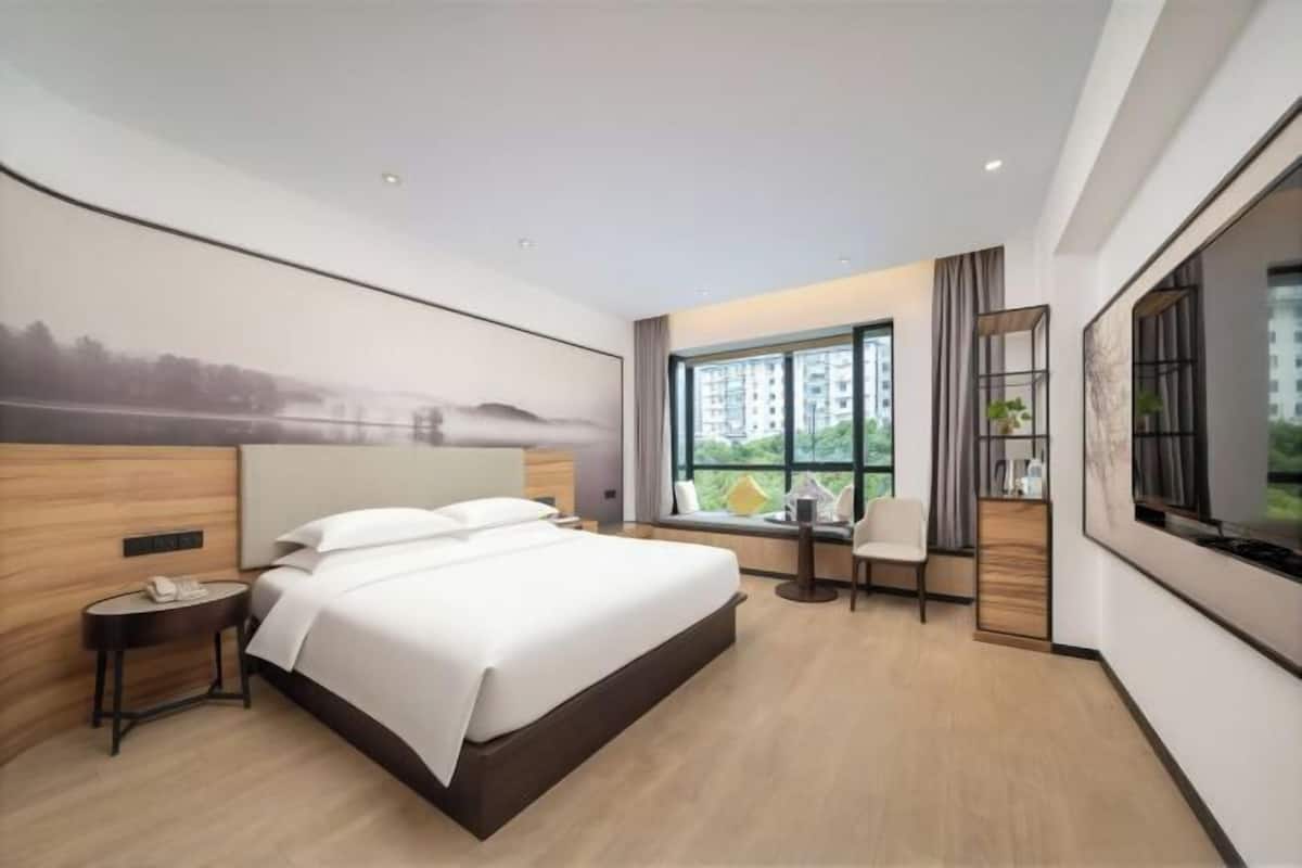 A modern hotel room with a large bed, a desk, and a view of greenery.