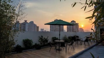 A rooftop terrace with a table and chairs, a green umbrella, and a city skyline at sunset.