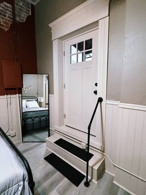 Une pièce avec une porte blanche, un lit doté d’une tête de lit noire, une petite fenêtre et un petit marchepied noir menant à la porte.