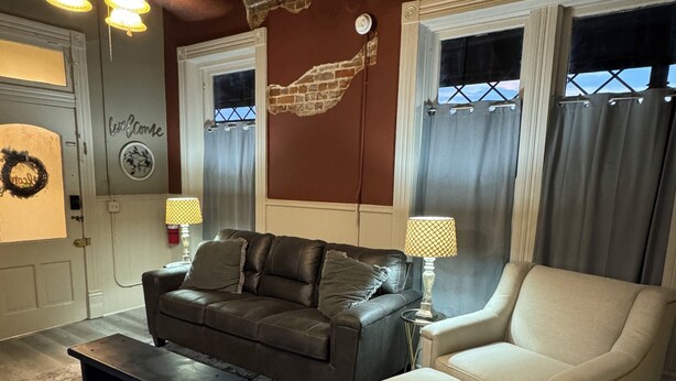 Ein Wohnzimmer mit einem braunen Ledersofa, einem beigefarbenen Sessel, einem Couchtisch und einem gemusterten Teppich. Zwei Lampen stehen auf Beistelltischen, ein Deckenventilator ist vorhanden, und eine Wand weist eine Ziegeloptik auf, mit einem Schild mit der Aufschrift 'Welcome'.