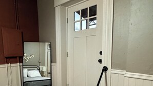 Ein Zimmer mit einer weißen Tür, einem Bett mit Kopfteil, einem kleinen Fenster und einer schwarzen Fußmatte.