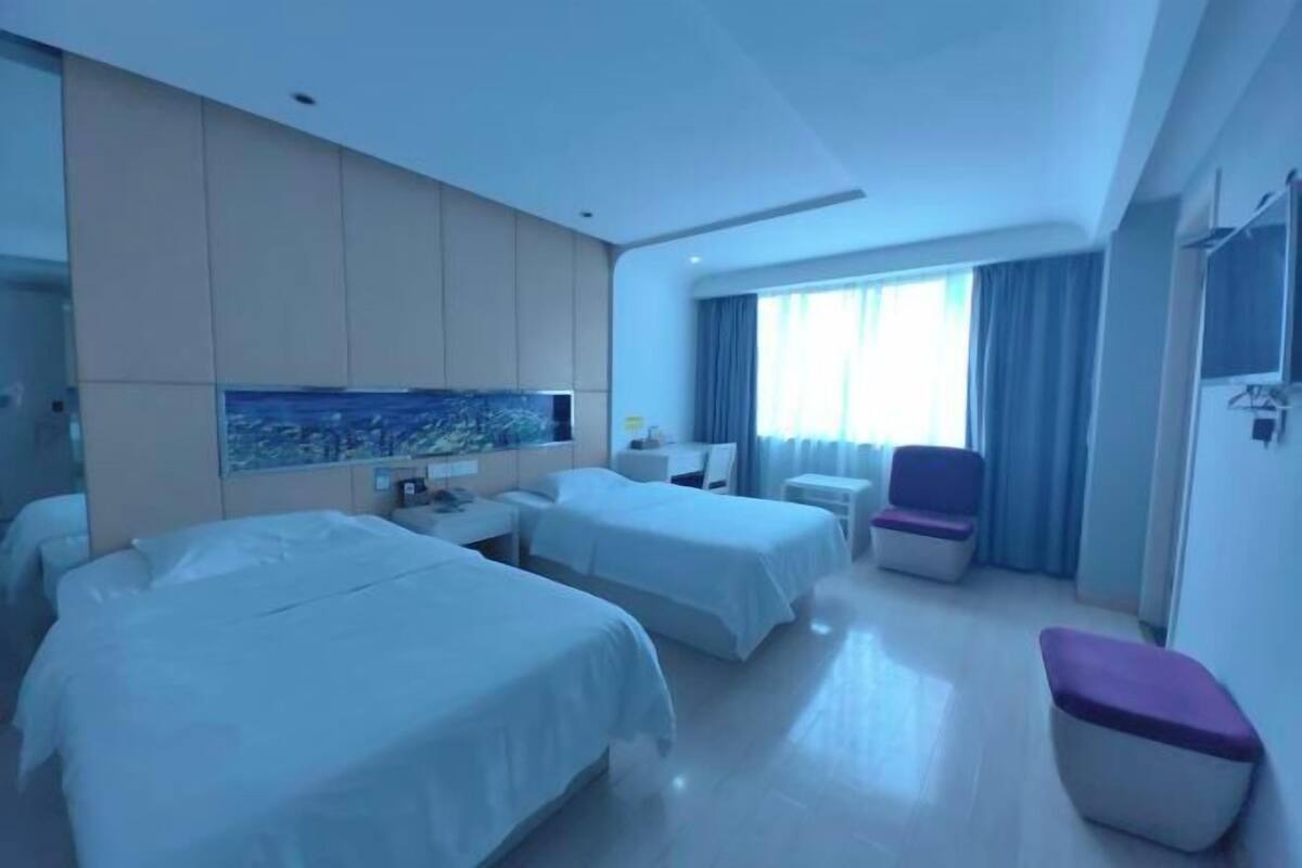 A hotel room with two beds, a small table, a chair, and a large window with curtains.