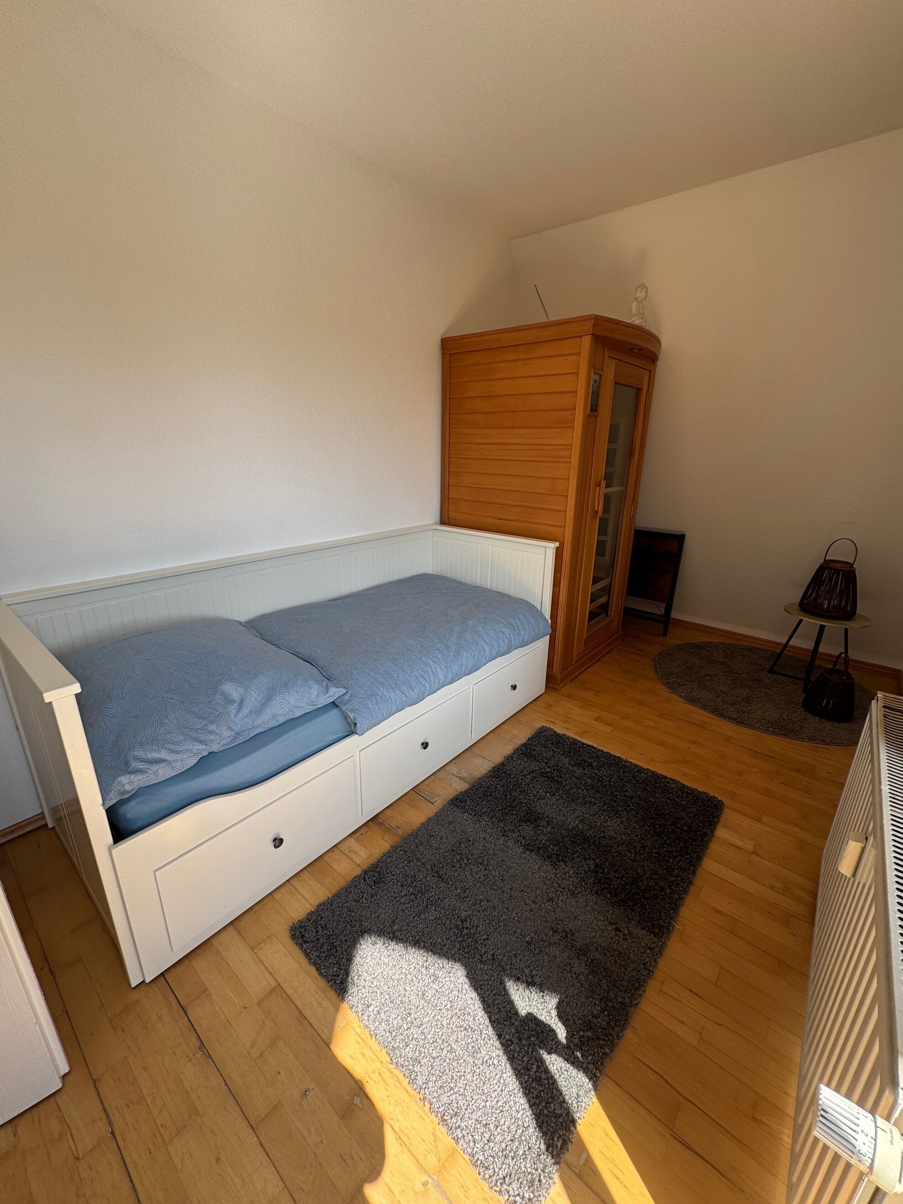 Een kleine eenpersoonskamer met een houten garderobe, een tafeltje en een radiator.